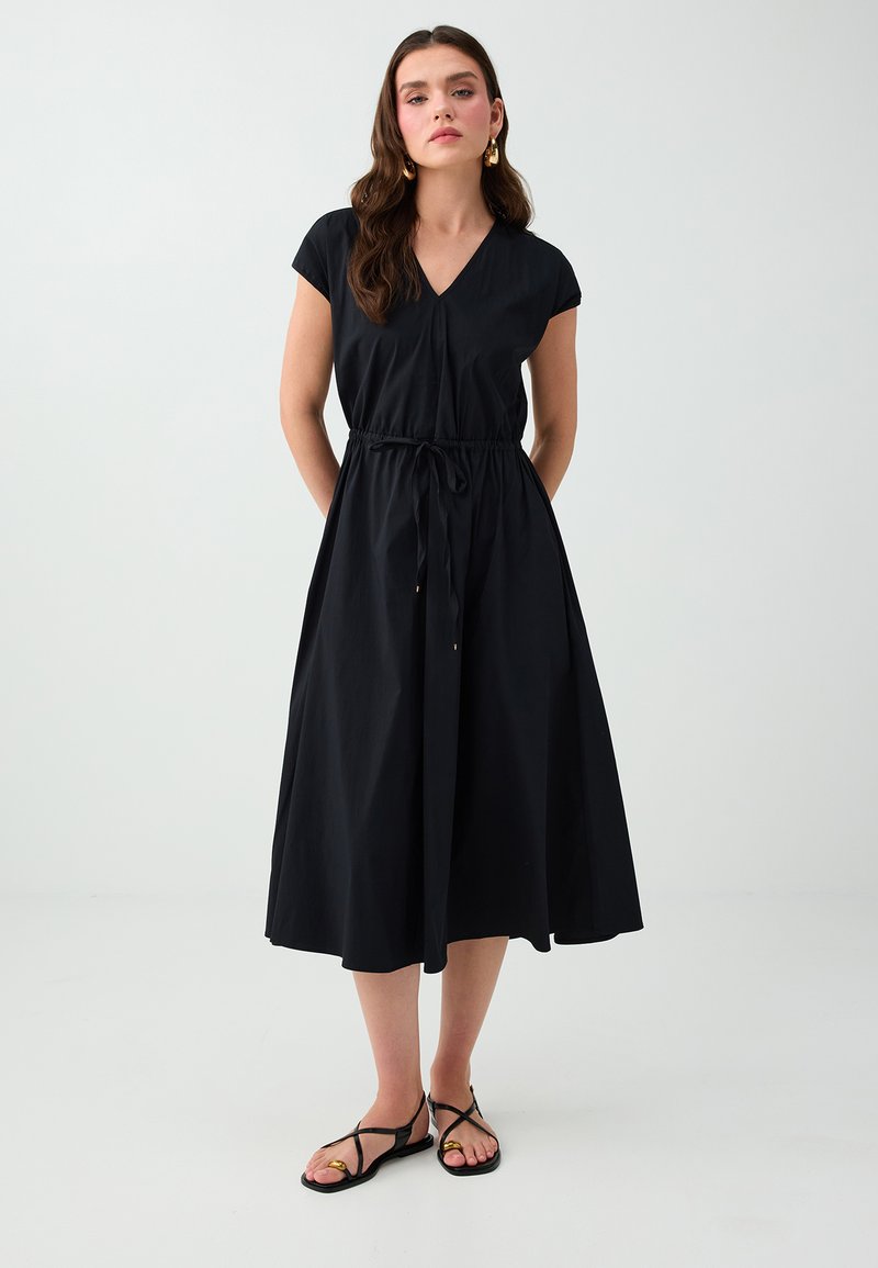Femme aux longs cheveux bruns porte une robe midi noire avec une taille à cordon et des sandales plates noires à brides sur un fond blanc uni.