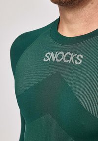T-shirt de sport vert en tissu texturé et respirant avec un motif en chevrons. Présente le nom de la marque "SNOCKS" en gris clair sur la poitrine.
