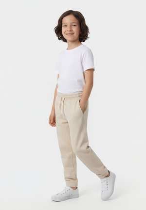 Enfant souriant aux cheveux bouclés portant un t-shirt blanc, un pantalon de survêtement beige avec cordon de serrage, et des baskets blanches, debout avec une main dans la poche.