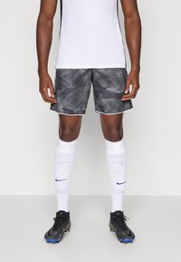 Mandlig atlet iført sorte mønstrede shorts, hvide sportsstrømper med skinnebeskyttere og sorte fodboldstøvler, der står på en ensfarvet baggrund.