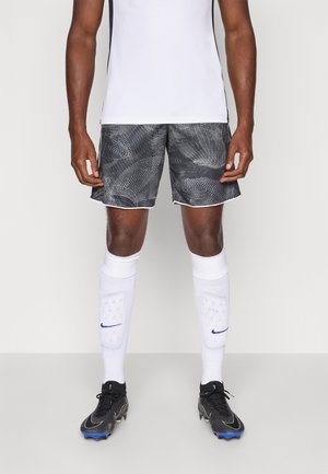 Männlicher Athlet in schwarzen gemusterten Shorts, weißen Sportsocken mit Schienbeinschonern und schwarzen Fußballschuhen, der vor einem einfarbigen Hintergrund steht.