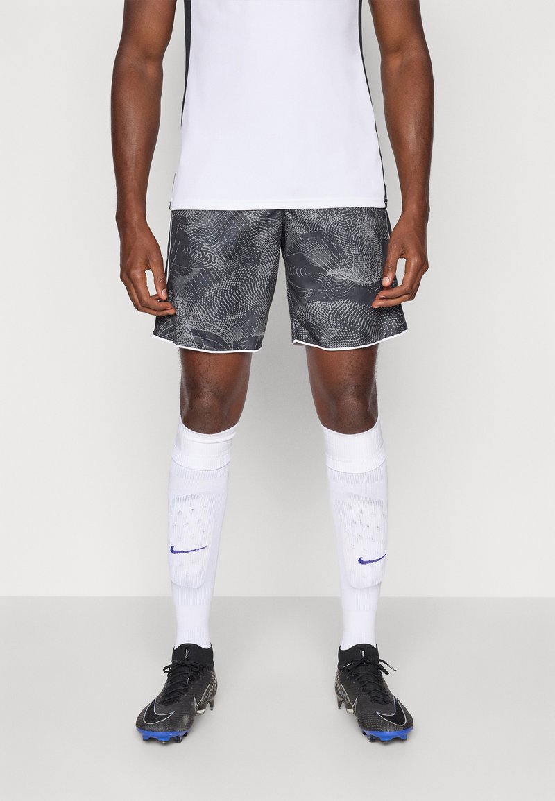 Mandlig atlet iført sorte mønstrede shorts, hvide sportsstrømper med skinnebeskyttere og sorte fodboldstøvler, der står på en ensfarvet baggrund.
