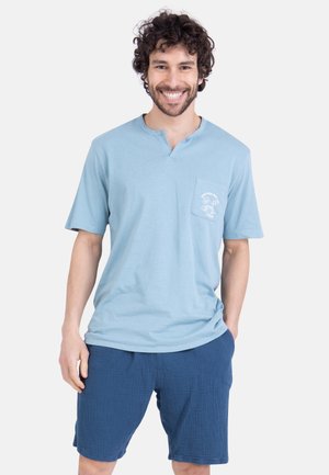 Homme souriant aux cheveux bouclés portant une chemise bleu clair à manches courtes avec poche poitrine et un short texturé bleu foncé, debout, détendu.