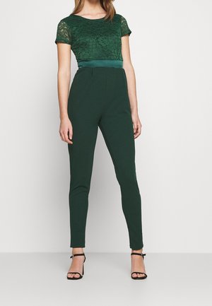 Femme portant un top en dentelle vert foncé à manches courtes et un pantalon ajusté vert foncé taille haute avec des talons noirs à bride à la cheville, debout sur un fond uni.