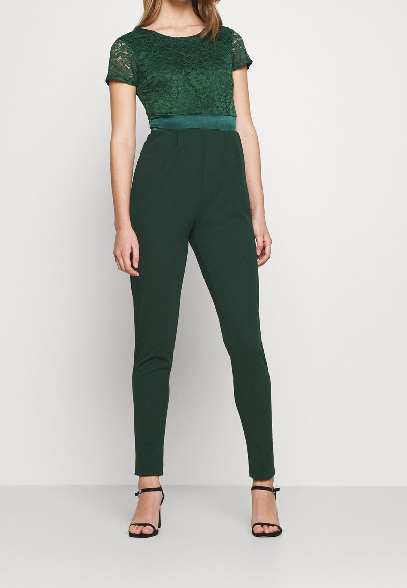 Femme portant un top en dentelle vert foncé à manches courtes et un pantalon ajusté vert foncé taille haute avec des talons noirs à bride à la cheville, debout sur un fond uni.