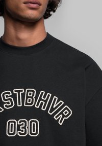 Černá mikina s kulatým výstřihem, s bílým vyšívaným nápisem "STBHVR 030" ve vyšitém designu. Měkká textilie s volným střihem.