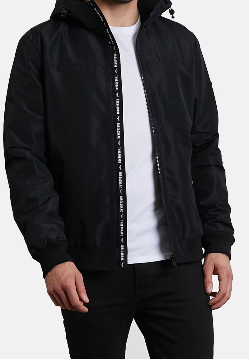 Homme portant une veste noire zippée sur une chemise blanche avec un pantalon noir, debout devant un fond clair uni.