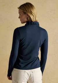 Mornarsko modra dolga majica z nabranim ovratnikom, mehke teksture, oprijetega kroja in minimalnim šivnim detajlom, v kombinaciji s svetlim hlačami.