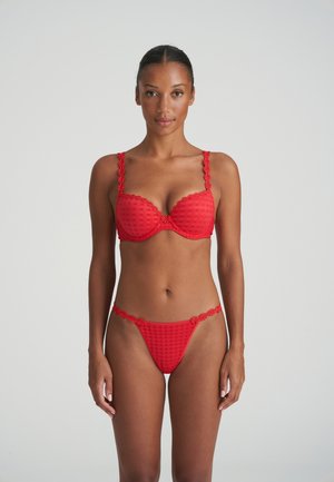 Mujer de pie mirando hacia adelante, vistiendo un conjunto de lencería roja con ribete floral y patrón a cuadros sobre un fondo liso y claro.