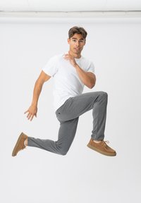 Grå sweatpants, hvid t-shirt og brune sko. Manden springer med det ene ben hævet og hænderne placeret i luften mod en ensfarvet baggrund.