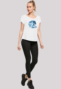 Hvit t-skjorte med korte ermer og grafikk av en blå hest, kombinert med svarte leggings og svarte joggesko. En enkel, avslappet passform.