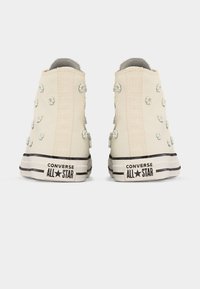 Αντιλευκά ψηλά παπούτσια Converse All Star με μικρές λευκές διακοσμήσεις σε σχήμα λουλουδιών, ορατά από πίσω σε απλό φόντο.
