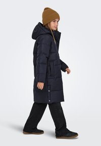 Temno modra puhovka z ruto, s kviltanim vzorcem, stranskimi žepi in sproščenim krojem. Nošena s črnimi hlačami in rjavo pleteno kapo.