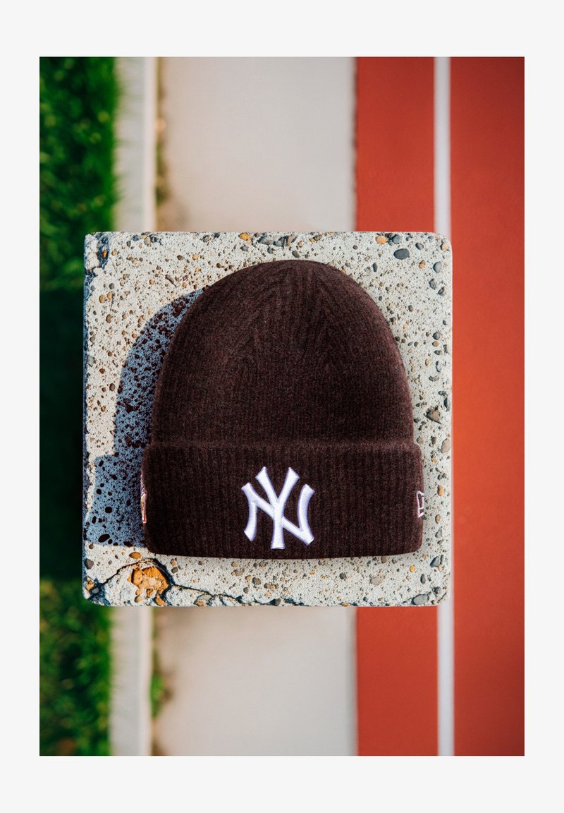 Gorro de malha castanho escuro com o logótipo branco "NY" descansando sobre um bloco de concreto texturizado, perto de relva verde e faixas de pista de corrida vermelhas.