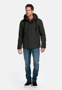 Donkergroene hoodiejas met voorzakken, ritssluiting en verstelbare koorden, gecombineerd met blauwe jeans en zwarte schoenen.