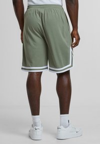 Olive green athletic shorts with white trim, elastic waistband, side pockets, and a sporty design. Worn with white sneakers and socks.