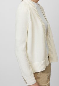 Persona che indossa un cardigan bianco a maglia sopra una camicia bianca, con pantaloni marrone chiaro, in piedi davanti a uno sfondo semplice.