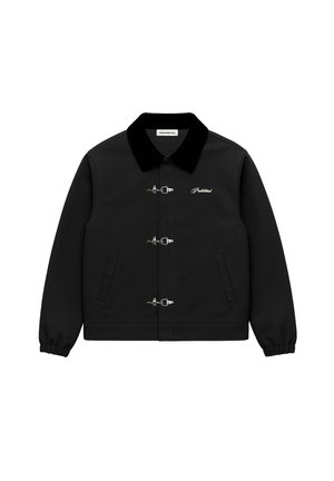 Mann, der eine rote Strickmütze und eine schwarze Jacke mit drei silbernen Knebelverschlüssen, ein rot kariertes Hemd und schwarze Hosen mit weißen Streifen trägt.