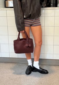 Woven burgundy handbag with two decorative pom-poms, sturdy handles, and a rectangular shape. Accompanied by black patent loafers and white socks.