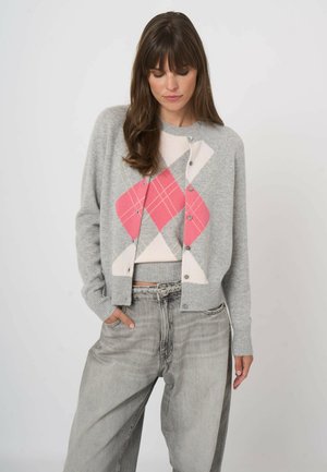 Kobieta w szarym kardiganie z różowo-białym wzorem w romby, w luźnych jasnych dżinsach, stoi z jedną ręką w kieszeni.