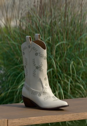 Creamfarget cowboystøvel med blomsterbroderi og middels brunt vinklet hæl, vist på en treoverflate med høyt gress i bakgrunnen.