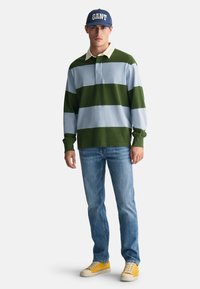 Green and light blue striped long-sleeve polo shirt with a white collar, paired with blue jeans and yellow sneakers, set against a white background.