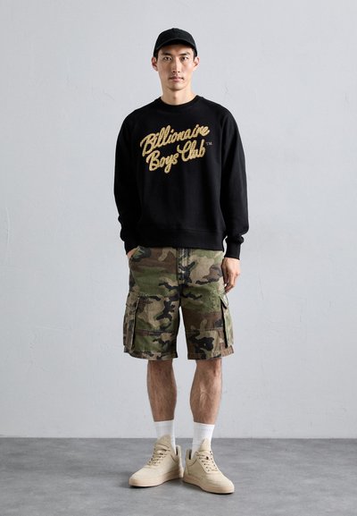 Melns sweatshirt ar zelta "Billionaire Boys Club" tekstu, kamuflāžas krāsu šorti, baltas zeķes un bežīgas kedas ar teksturētu apdari.