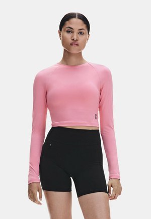 Donna che indossa un crop top rosa aderente a maniche lunghe e pantaloncini sportivi neri a vita alta, in piedi davanti a uno sfondo semplice.