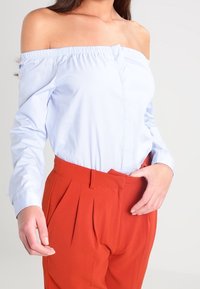 Světlomodrá halenka odhalená na ramenou s elastickým výstřihem a zapínáním na jeden knoflík, v kombinaci s červenými kalhotami s vysokým pasem a sklady.