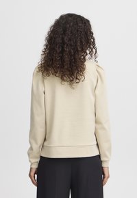 Beige Sweatshirt mit langen Ärmeln, strukturiertem Stoff und gerafften Details an den Schultern; taillierter Bund; getragen mit schwarzen Hosen.