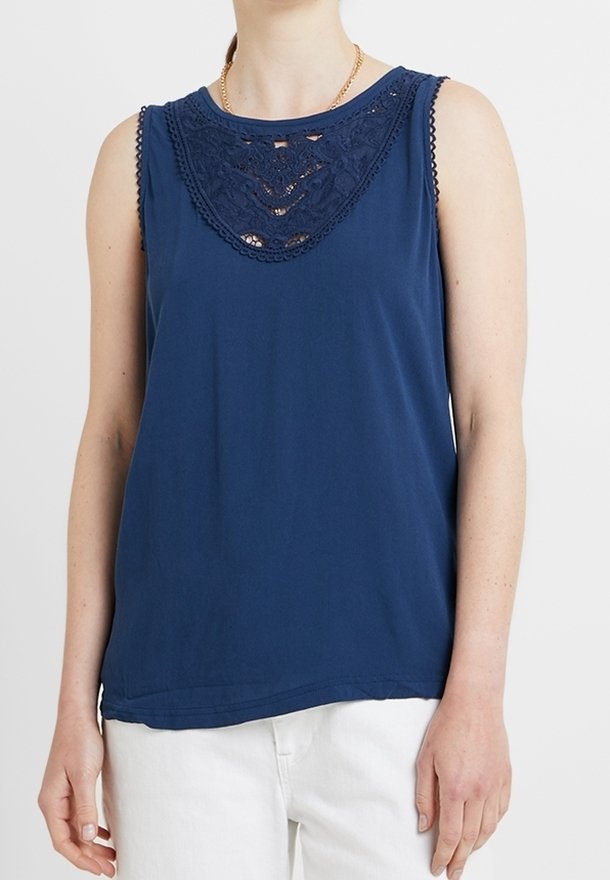 Femme portant un haut sans manches bleu marine avec un détail en dentelle au niveau du col et un pantalon blanc sur un fond uni clair.