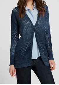 Kobieta w granatowym kardiganie o otwartym splotem, nosząca jasnoniebieską bluzkę i ciemne dżinsy, stoi na tle jednolitego tła.