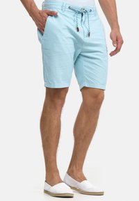 Mann, der hellblaue Shorts mit Kordelzug und weiße Slip-on-Schuhe trägt, steht mit einer Hand in der Tasche vor weißem Hintergrund.