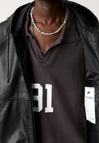 Uomo che indossa una giacca di pelle nera sopra una camicia scura con colletto e la stampa bianca "81", accompagnato da due collane, una con perline colorate e una con una catena d'argento.