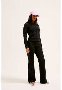 Veste de sport noire zippée associée à un pantalon évasé assorti. Le modèle porte une casquette rose clair. Les chaussures sont des sandales à bout ouvert.