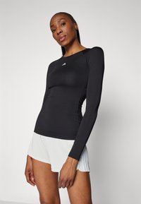 Haut de sport noir à manches longues avec un col rond et une coupe ajustée. Présente un petit logo blanc sur la poitrine. Style simple, tissu extensible.