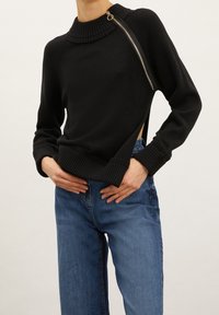 Svart tröja med hög ribbad krage, zip-detalj på sidan och öppen midja. Matchad med avslappnat passande blå denimjeans.