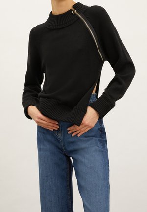 Zwarte trui met een hoge geribbelde kraag, zijritsdetail en open taille ontwerp. Gecombineerd met relaxte blauwe denim jeans.