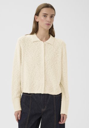 Ženska, oblečena v kremasto teksturiran pulover s tipkami in ovratnikom ter temne kavbojke z visokim pasom, stoji ob belem svetlem ozadju.