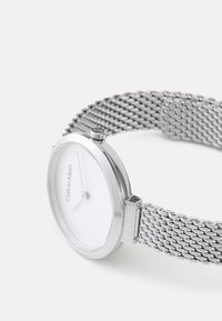 Montre avec un bracelet en mesh argenté, un cadran blanc rond, un design minimaliste et un logo Calvin Klein sur le cadran. Équipée d'une boucle en métal.