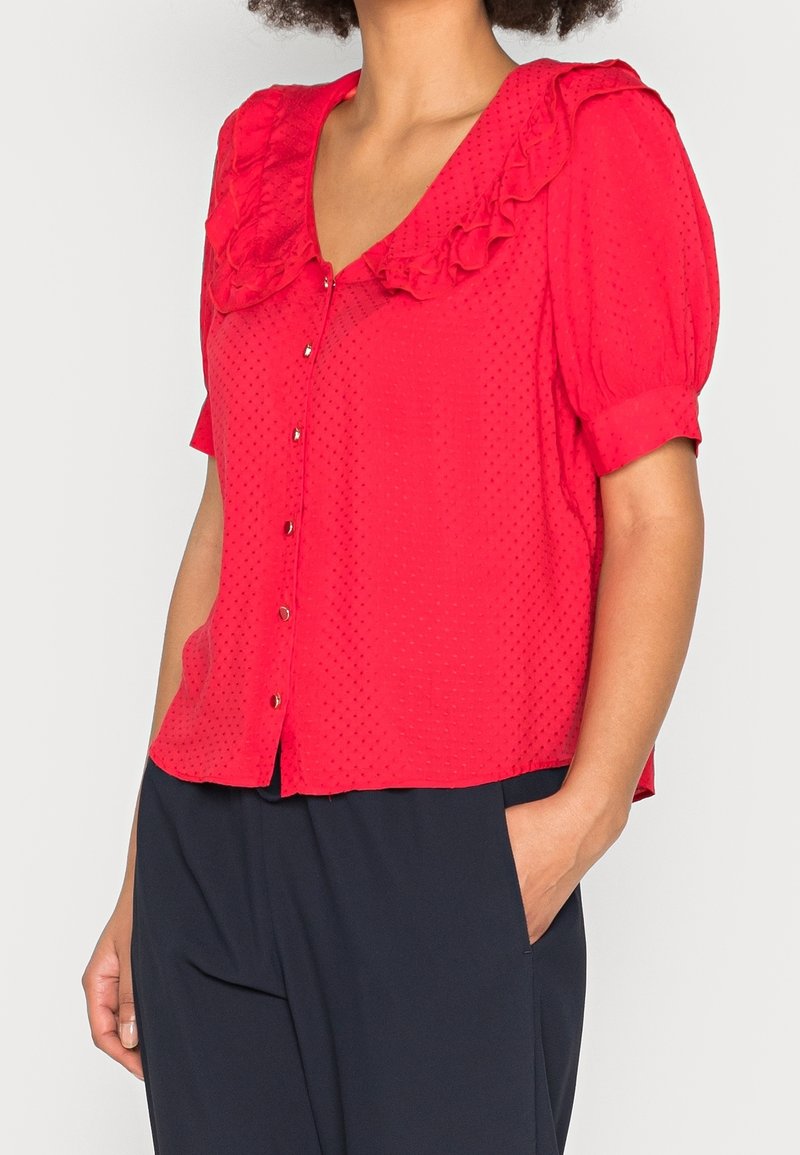 Blusa rossa con scollo arricciato e maniche corte a sbuffo, dotata di chiusura frontale con bottoni e tessuto testurizzato con piccole decorazioni a pois.