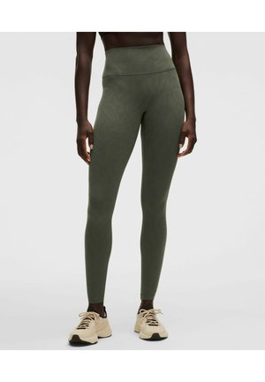Frau, die hoch taillierte olivgrüne Leggings und beige Turnschuhe trägt, vor einem schlichten hellen Hintergrund stehend.