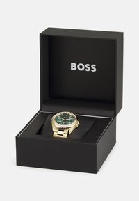Montre en or avec un cadran vert, trois sous-cadrans et une lunette accentuée de cristaux, présentée dans une boîte noire avec le logo "BOSS".