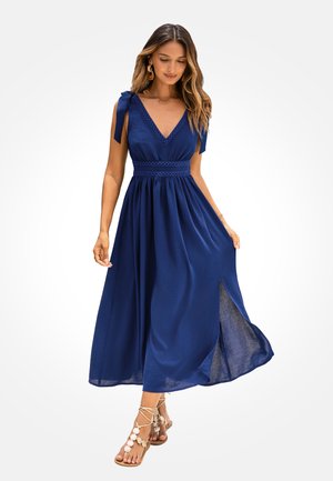 Woman wearing a flowy navy blue sleeveless dress with shoulder ties, waist detailing, side slit, and beige lace-up sandals with shell embellishments.