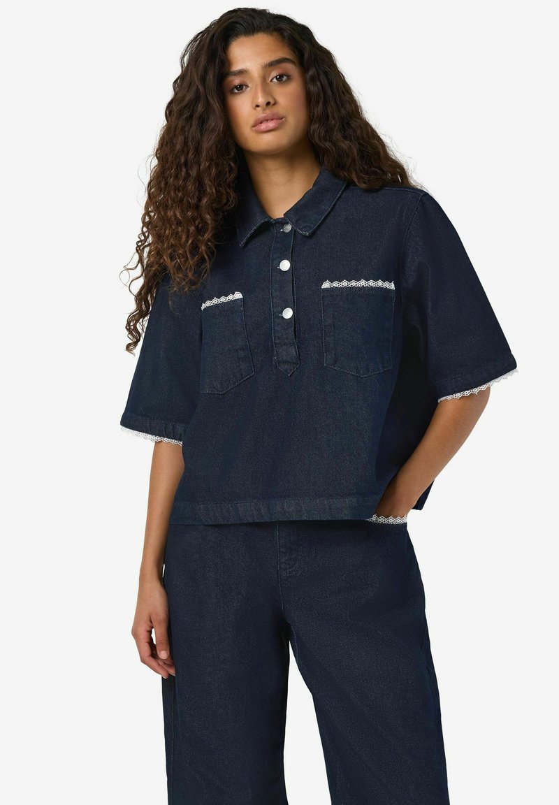 Temno denim kratka srajca z belim čipkastim robom na rokavih in žepih. Ima ovratnik, sprednjo zapenjanje na gumbe in dve prsni žepi.