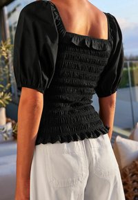 Černý top s nafouknutými rukávy a smockovaným tělem, doplněný bílými kalhotami. Design obsahuje čtvercový výstřih.