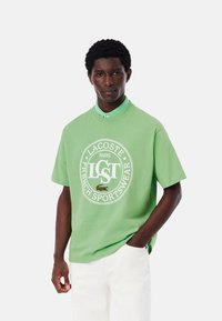 Camiseta de algodón verde con un logo redondo que presenta "LACOSTE PARIS FRENCH SPORTSWEAR" en blanco. Mangas cortas, ajuste relajado, combinada con pantalones blancos.