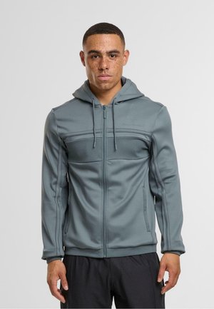 Sudadera gris con cremallera y un panel superior texturizado, detalles de malla en los laterales y capucha con cordón. Combinada con pantalones cortos deportivos negros.