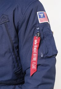 Mornarji modra pilotska jakna z značko z ameriško zastavo, zadrgo na žepu in rdečim oznako "ODSTRANITE PRED LETOM".