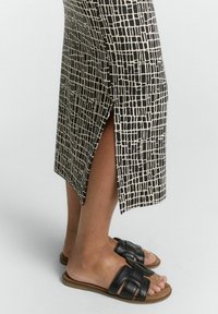 Jupe midi à motifs noirs et beiges avec fente sur le côté. Associée à des sandales à glisser en cuir noir avec un design de bande tressée.